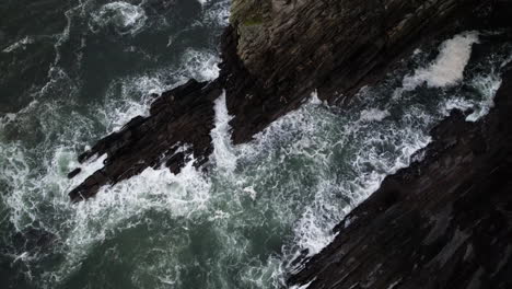 Birds-Eye-Aerial-View,-Ocean-Waves-Crushing-Under-Steep-Rocky-Cliffs-of-Ireland