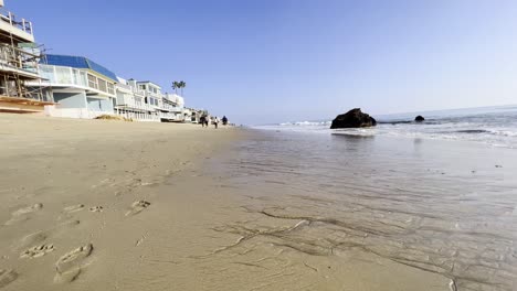 People-walking-at-the-beach-in-Malibu,-homes-facing-the-ocean-and-waves-rolling