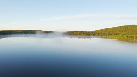 Aerial-view-of-blue-foggy-lake-in-sunrise-with-golden-hour-green-forest-landscape-in-the-background-filmed-in-Lapland-Finland