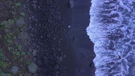 Man-Walking-Along-Waves-Swashing-On-Volcanic-Dark-Sand-Beach,-Aerial