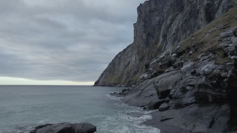 Aerial-drone-view-low-over-the-rocky-coast,-under-steep-mountains,-gloomy-evening,-in-Lofoten,-Norway