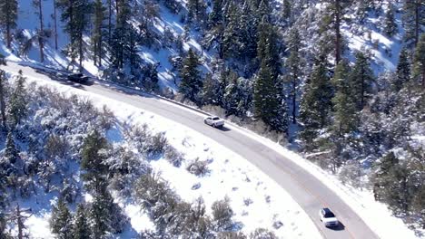 Slippery,-snowing-mountain-road-with-two-way-traffic-traveling-along-the-winding-alpine-route-through-a-pine-forest---scenic-aerial-view