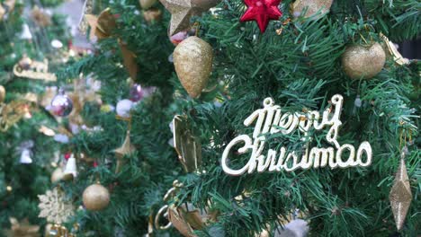 Zahlreiche-Dekorationsornamente-Und-Die-Aufschrift-„frohe-Weihnachten“-Hängt-An-Einem-Grünen-Weihnachtsbaum,-Während-Eine-Maschine-In-Hongkong-Falsche-Schneeflocken-Abfeuert