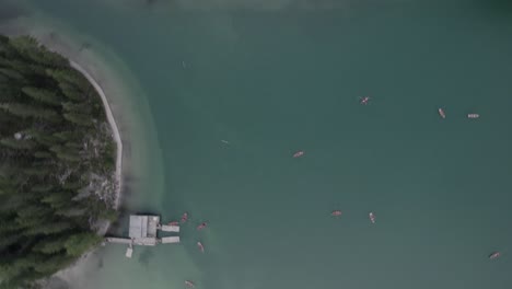 Drone-video-with-a-descending-cenital-plane-in-a-spiral-shape,-brand-of-the-house,-over-the-lake-of-braies-with-boats-with-people-rowing