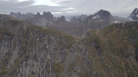 Aerial-drone-view-towards-a-steep-mountain-wall-with-alpine-peaks-in-the-background,-in-Lofoten,-Norway