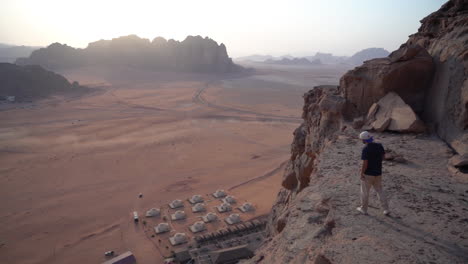 Man-Walking-on-Cliff-Above-Bedouin-Camp-and-Desert-Landscape-of-Wadi-Rum,-Jordan-With-Misty-Horizon,-Slow-Motion