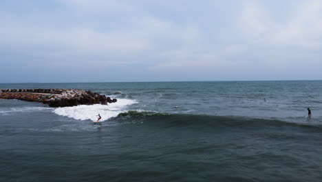 Aerial,-person-surfing-on-ocean-wave-in-a-bay-protected-by-a-tetrapod-seawall