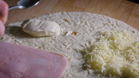 A-close-up-shot-of-a-dollop-of-rich-creamy-mayonnaise-being-added-to-a-ham-and-cheese-tortilla-wrap,-the-condiment-being-spread-evenly-using-the-back-of-a-spoon
