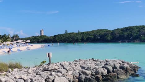 Pan-shot-of-trees,-rocks-and-a-sandy-beach-with-umbrellas-sunchairs-and-people-on-a-sunny-bright-day-in-The-Bahamas,-Blue-Lagoon-Island-|-4K-30-FPS