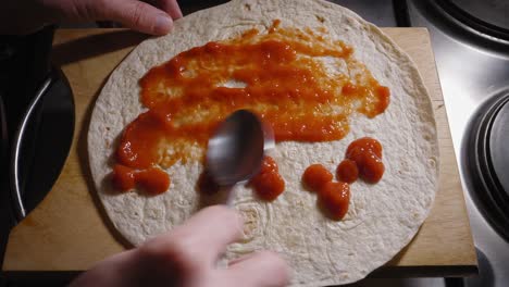 A-top-down-shot-of-the-hands-of-a-chef-evenly-spreading-a-tasty-tomato-source-around-a-tortilla-wrap-with-the-back-of-a-spoon,-the-source-providing-a-layer-of-flavour-to-a-delicious-Hot-Dog-tortilla