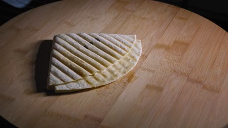 A-close-up-shot-of-a-freshly-grilled-Quesadilla-being-placed-onto-a-cutting-board,-oil-from-the-melted-cheese-dripping-off-the-toasted-tortilla-wrap-onto-the-wooden-surface