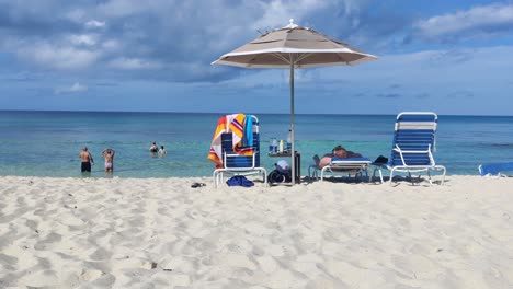 Menschen-Im-Meer-Genießen-Den-Heißen-Sommertag-Auf-Der-Blauen-Laguneninsel-Auf-Den-Bahamas-Mit-Sonnenliege-Und-Regenschirm,-Handheld-rückansicht,-4k-30-Fps