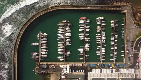 Aerial-top-down-sail-boat-moored-in-harbor,-drone-eye’s-bird-view-of-concrete-port-with-ocean-waves-crashing