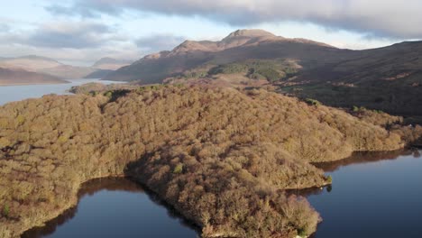 Rising-and-panning-drone-footage-of-an-autumn-sunrise-over-Loch-Lomond-and-Ben-Lomond-mountain-in-the-Loch-Lomond-and-the-Trossachs-National-Park,-Scotland-with-native-broadleaf-woodland-on-the-shore