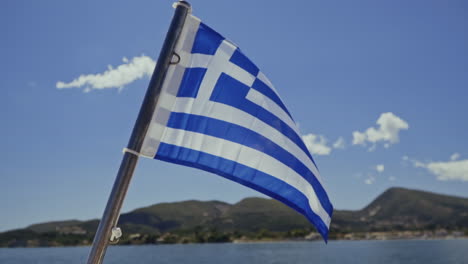National-flag-of-Greece-blows-in-the-wind-on-boat-with-sea-and-coast-behind