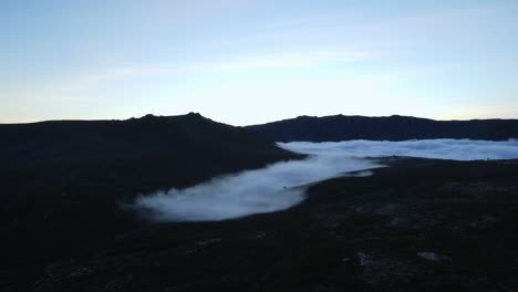 Aerial-far-away-view-of-a-valley-covered-with-fog-and-clouds-drifting-away
