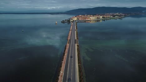 Cars-traffic-on-bridge-road-across-the-lagoon-toward-the-island-town-Orbetello-close-to-Monte-Argentario-and-the-Maremma-Park-in-Tuscany,-Italy,-with-blue-sky-and-calm-blue-water