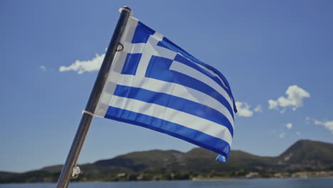 Bandera-Griega-Ondeando-En-El-Viento-En-Un-Barco-Con-Una-Isla-Mediterránea-Detrás