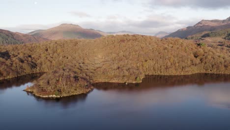 Aerial-drone-footage-of-an-autumn-sunrise-over-the-mountains-Ben-Lomond-and-Beinn-Bhreac-in-Loch-Lomond-and-the-Trossachs-National-Park,-Scotland-with-native-broadleaf-woodland-on-Loch-Lomond-shore