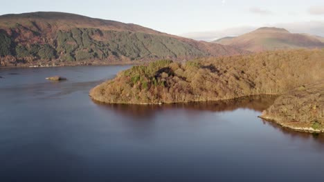 Left-roll-with-righ-pan-aerial-drone-footage-revealing-Ben-Lomond,-Beinn-Dubh-and-Beinn-Bhreac-mountains-during-an-autumn-sunrise-with-native-broadleaf-woodland-and-Loch-Lomond-foreground