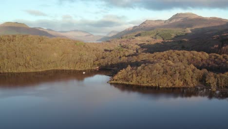 Left-roll-aerial-drone-footage-of-Ben-Lomond-and-Beinn-Bhreac-mountains-during-an-autumn-sunrise-with-native-broadleaf-woodland-and-Loch-Lomond-in-Loch-Lomond-and-the-Trossachs-National-Park
