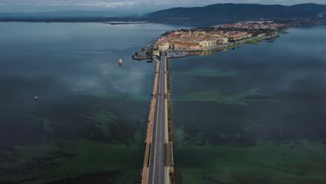 Cars-on-bridge-traffic-road-across-the-lagoon-toward-the-island-town-Orbetello-close-to-Monte-Argentario-and-the-Maremma-Park-in-Tuscany,-Italy,-with-blue-sky-and-calm-blue-water