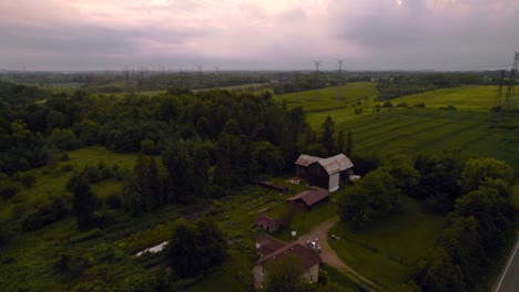 Cinematic-Reveal-of-Farm-House-next-to-a-Rural-Country-Roadway-with-Farmer's-Field-and-Power-lines-under-cloudy-skies-at-Sunset