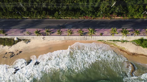 Slow-motion-top-view-of-the-beautiful-landscape-of-the-beach-water-waves-crashing-to-the-sand-next-to-the-coastal-road-on-the-sunset-of-Vietnam,-mui-ne-at-palm-trees-forest-background-of-the-road