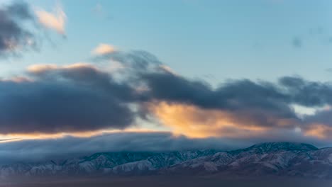 Light-dusting-of-snow-on-the-Mojave-Desert-mountains-with-sunset-clouds-rolling-over-the-rugged-peaks---time-lapse