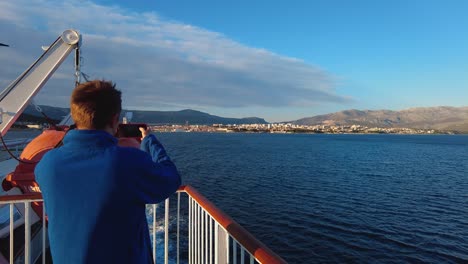 A-tourist-recording-a-video-on-his-phone-of-coastline-in-open-water-while-the-boat-is-sailing-from-Split-to-island-Vis-in-Croatia,-in-the-Adriatic-Sea