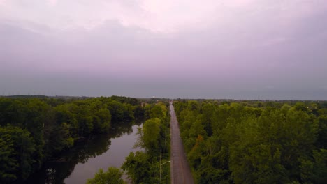 Aerial-Drone-Tracking-shot-of-Country-Road-at-Dusk-Next-to-Pond-and-Forested-area-with-Cloudy-Skies-scenic-Landscape
