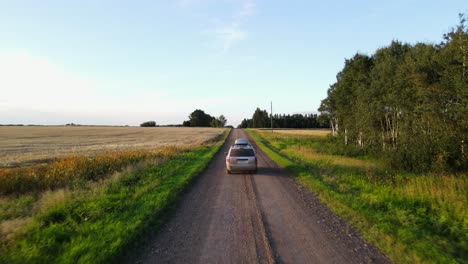 Aerial-tracking-shot-of-a-silver-minivan-driving-along-a-dusty-dirt-road-during-sunset-in-the-Canadian-countryside