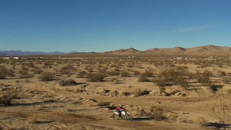 Riding-a-motorcycle-along-a-bumpy-dirt-trail-in-the-Mojave-Desert-and-jumping-across-a-road-in-slow-motion---aerial-view