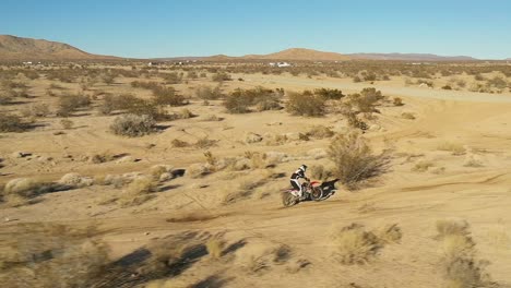 Motorcyclist-riding-along-a-bumpy-dirt-trail-in-the-Mojave-Desert-and-jumping-a-road-in-slow-motion---aerial-view