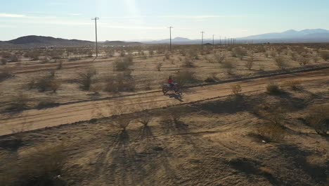 Motorcycle-rider-turning-off-a-dirt-road-onto-a-trail-on-a-Mojave-Desert-off-road-adventure---aerial-view-in-slow-motion