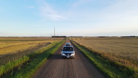 Backwards-moving-aerial-tracking-shot-of-a-silver-mini-van-driving-along-a-dusty-dirt-road-through-the-countryside-of-central-Alberta