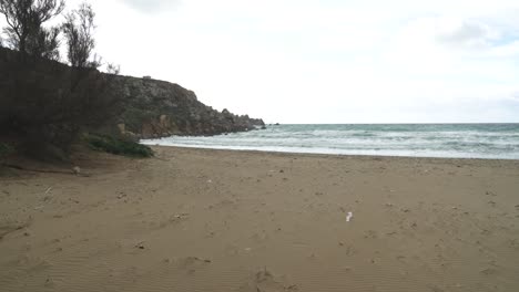 Caminando-Lentamente-En-La-Playa-De-Arena-Dorada-De-La-Bahía-En-Un-Día-Nublado-Con-El-Rugiente-Mar-Mediterráneo-En-Malta