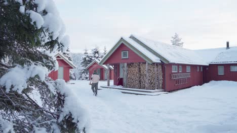 Mann-Mit-Seinem-Hund,-Der-Im-Winter-In-Norwegen-Auf-Dem-Schneebedeckten-Boden-Mit-Traditionellen-Hütten-Spazieren-Geht