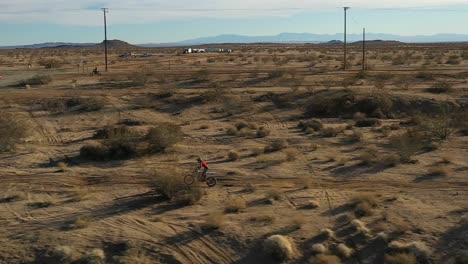Offroad-Motorradfahrer,-Der-Auf-Einem-Holprigen-Pfad-In-Der-Mojave-Wüste-Fährt-Und-Dann-Auf-Eine-Unbefestigte-Straße-Springt---Luftbild-In-Zeitlupe