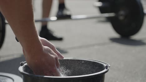 Close-up-of-young-man-puts-chalk-on-his-hands-from-bucket-and-claps-so-the-air-fills-with-dust-before-lifting-wait-at-cross-fit-outside-in-4k-slow-motion