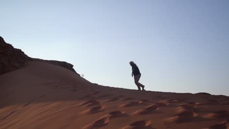 Silhouette-of-Man-Walking-Uphill-on-Dune-Sand-in-Desert-Landscape-on-Hot-Sunny-Evening