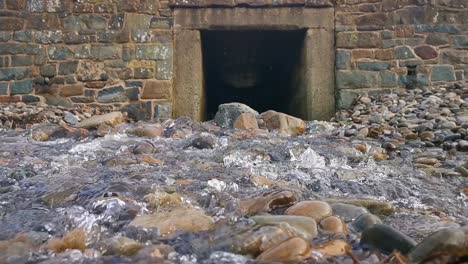 Water-cascading-from-a-hole-in-a-stone-wall-across-a-stony-beach-in-Broad-Haven,-West-Wales