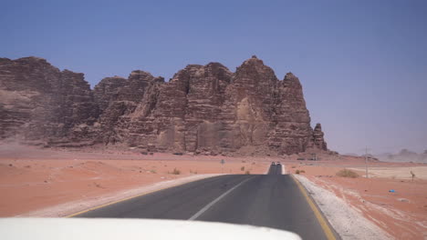Fahren-In-Der-Wüste-Auf-Asphaltstraße-Mit-Blick-Auf-Sandsteinfelsen-Und-Trockene-Landschaft-An-Heißen-Sonnigen-Tag,-Passagier-Pov,-Wadi-Rum-Jordan
