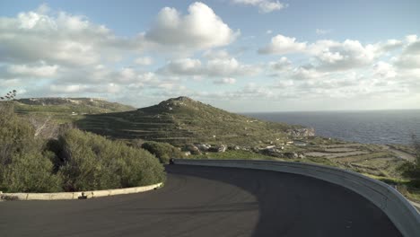 Curvy-Asphalt-Road-Leading-Down-Near-Coastline-of-Malta-with-Vast-Greenery-on-Plateu