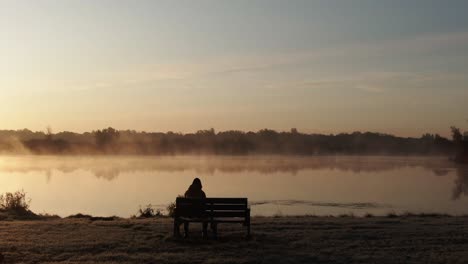 Back-view-of-a-lonely-person-sitting-on-a-bench-in-front-of-a-misty-lake-during-sunset,-sad-and-depressing-concept