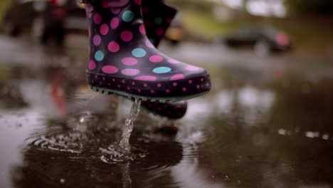 Unrecognizable-girl-jumping-in-a-pool-of-water-during-rainy-day-with-waterproof-boots,-close-up-of-the-feet-with-selective-focus