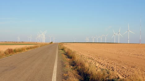 Una-Carretera-Asfaltada-Vacía-Entre-Campos-Con-Turbinas-Eólicas-Durante-El-Verano-En-Aragón,-España