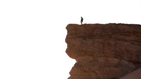 Man-walking-on-steep-sandstone-cliff-in-a-desert-with-sunlight-in-back-wide-view