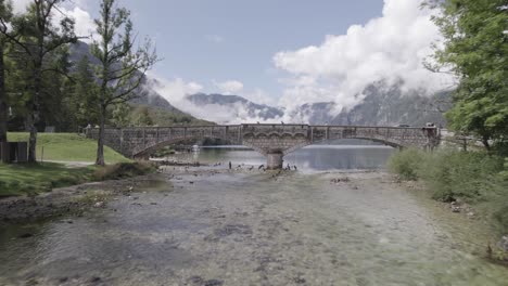 Frontal-drone-video-over-Lake-Bohinj-advancing-towards-Most-bridge-flying-over-the-river-level,-a-canoe-and-cyclists-can-be-seen-passing-the-bridge