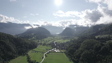 Frontal-drone-video-moving-over-farms-in-the-Triglav-with-hills-on-the-horizon,-the-sun-in-front-and-clear-sky-with-clouds